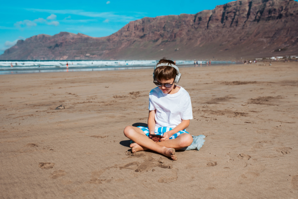 Boy watching video on smartphone with headphones on head. Young boy is online during family vacation in resort. Concept of a beach summer vacation with kids.