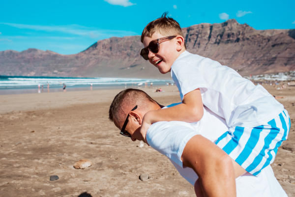 Father carrying son on back at the beach. Dad and boy having fun, enjoying sandy beach in Canary Islands. Concept of beach summer vacation with kids.