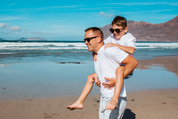 Father carrying son on back at the beach. Dad and boy having fun, enjoying sandy beach in Canary Islands. Concept of beach summer vacation with kids.