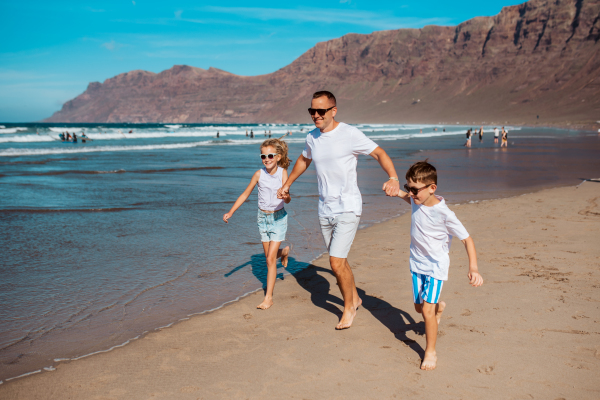 Father with two kids running on beach. Young family enjoying sandy beach, looking at crystalline sea in Canary Islands. Concept of a beach summer vacation with kids.