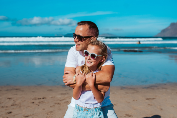 Father embracing daughter, standing on beach. Dad and girl enjoying sandy beach, looking at crystalline sea in Canary Islands. Concept of beach summer vacation with kids.