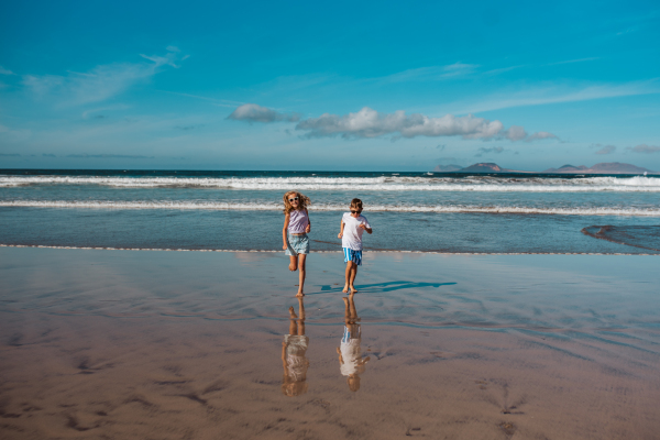 Siblings playing on beach, running, skipping in water. Smilling girl and boy on sandy beach of Canary islands. Concept of a family beach summer vacation with kids.