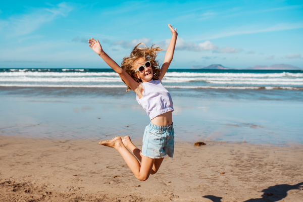 Jumping girl on the beach. Smilling blonde girl enjoying sandy beach, looking at crystalline sea in Canary Islands. Concept of beach summer vacation with kids.