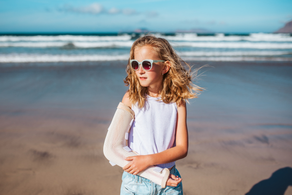 Girl with broken arm on beach. Young blonde girl has arm cast, injured during family vacation in holiday resort. Concept of a beach summer vacation with kids.