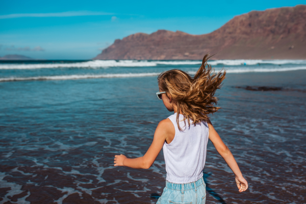 Rear view of girl on the beach. Blonde girl enjoying sandy beach, looking at crystalline sea in Canary Islands. Concept of beach summer vacation with kids.