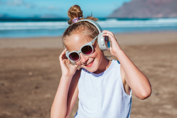 Girl with headphones on head standing on beach. Smilling blonde girl enjoying sandy beach, listening music via wireless headphones. Concept of beach summer vacation with kids.