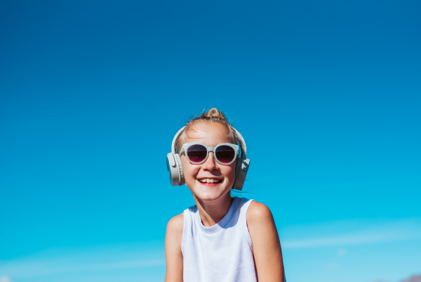 Girl with headphones on head standing on beach. Smilling blonde girl enjoying sandy beach, listening music via wireless headphones. Concept of beach summer vacation with kids.