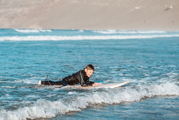 Young boy surfing in sea waves. Smilling boy in neoprene wetsuit lying on sufrboard. Concept of popular water sport for kids.