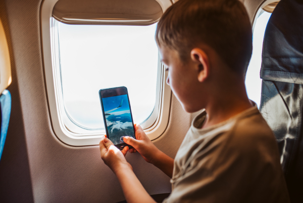 Boy with headphones sitting airplane, taking photos from window with smartphone. Concept of a famiily beach summer vacation with kids.