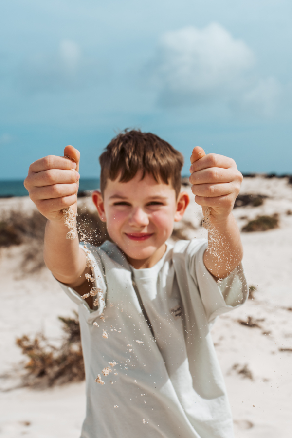 Boy with sand falling from his clnched fist, standing on beach. Family summer vacation by the sea.