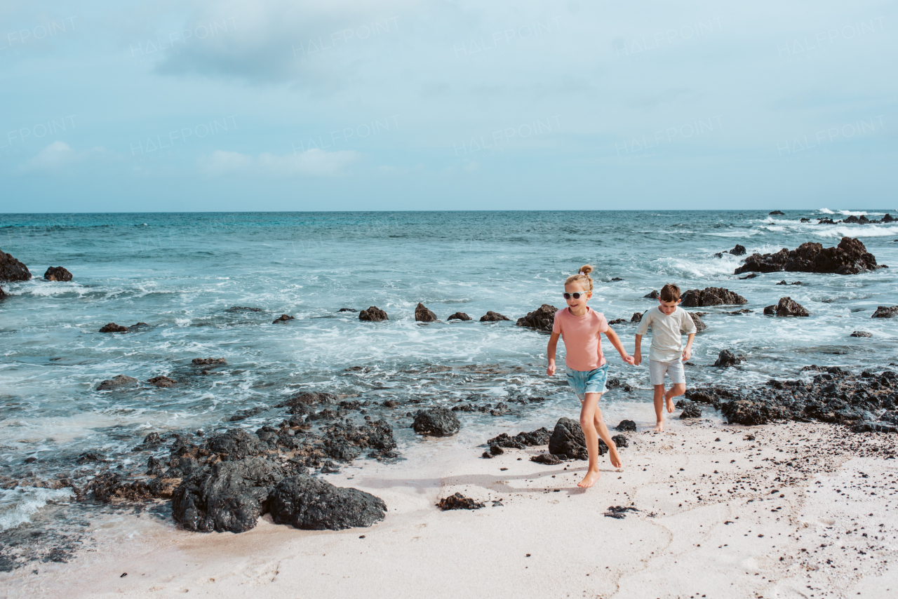 Siblings playing on beach, running, skipping rocks. Smilling girl and boy ion sandy beach with volcanic rocks of Canary islands. Concept of a family beach summer vacation with kids.