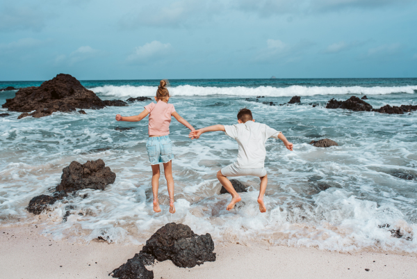 Siblings playing on beach, running, skipping rocks. Smilling girl and boy ion sandy beach with volcanic rocks of Canary islands. Concept of a family beach summer vacation with kids.