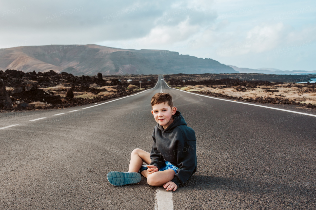 Portrait of boy sitting on empty road, looking at camera, smiling.