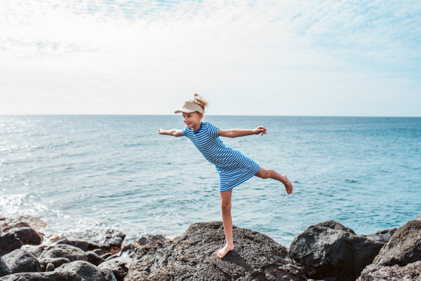 Young girl standing on one leg on rock. Blonde girl in dress and cap, visor enjoying vacation in Canary Islands. Concept of beach summer vacation with kids.