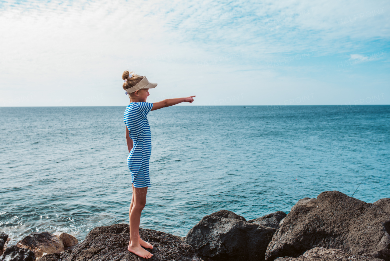 Side view of girl pointing at someting on sea. Blonde girl in striped dress enjoying vacation in Canary Islands. Concept of beach summer vacation with kids.