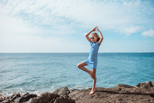 Young girl standing on one leg on rock. Blonde girl in striped dress enjoying vacation in Canary Islands. Concept of beach summer vacation with kids.