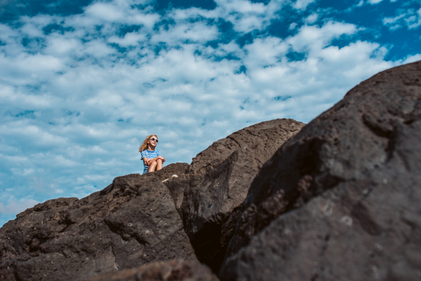 Portrait of girl sitting on rocks by sea.Blonde girl in striped dress enjoying vacation in Canary Islands. Concept of beach summer vacation with kids.