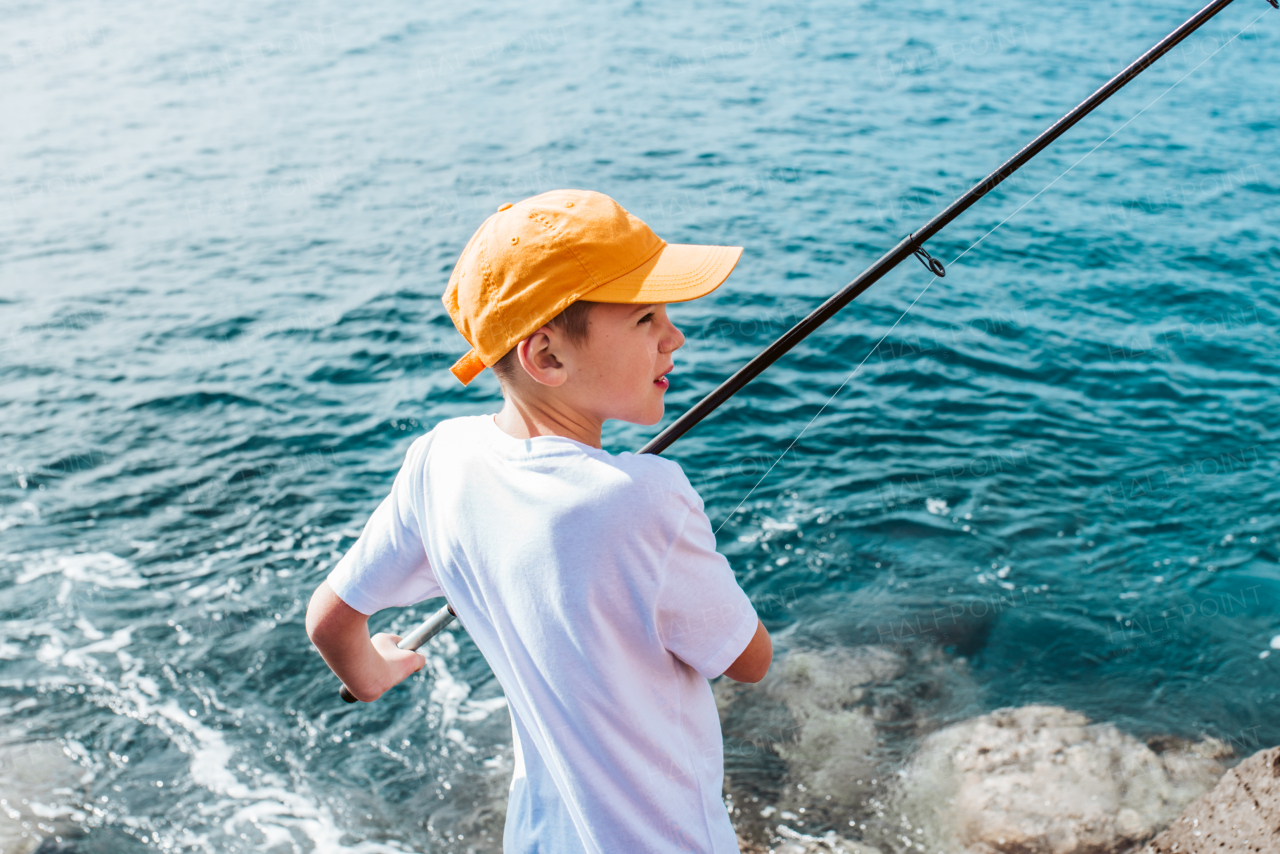 Young boy with cap on head fishing in the sea. Standing on rock, coast holding fish rod. Top view.