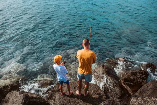 Father teaching young son fishing in the sea. Standing on rock, coast together holding fish rod. Top view of dad and boy.