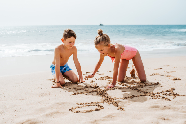 Siblings playing on beach, drawing tic-tac-toe in the sand, Smilling girl and boy in swimsuit on sandy beach of Canary islands. Concept of family beach summer vacation with kids.