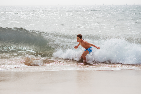 Young boy playing, running and splashing in strong sea waves. Smilling boy in swimsuit looking at waves. Concept of a beach summer vacation with kids.