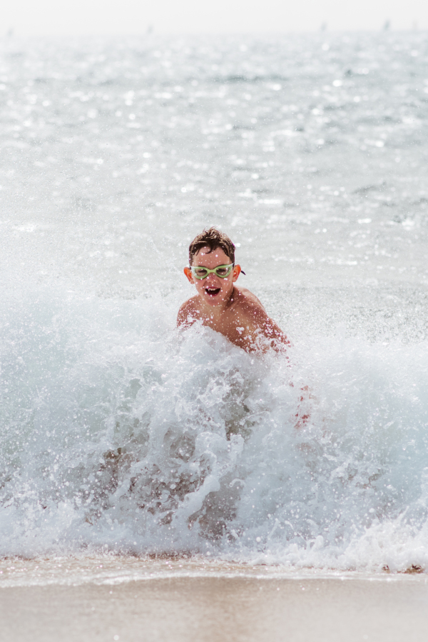 Young boy playing, running and splashing in strong sea waves. Smilling boy in swimsuit looking at waves. Concept of a beach summer vacation with kids.