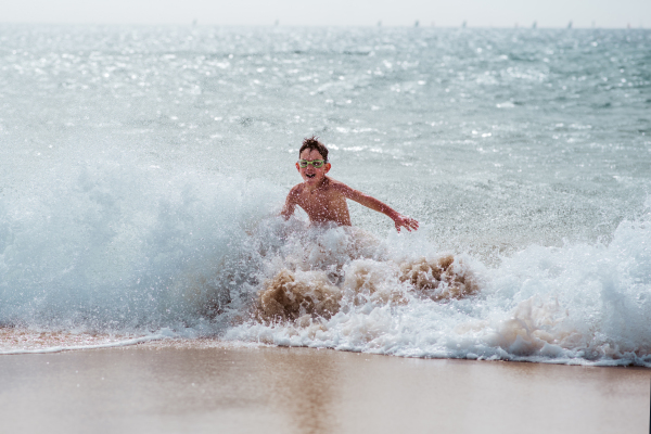 Young boy playing, running and splashing in strong sea waves. Smilling boy in swimsuit and swimming goggles. Concept of a beach summer vacation with kids.