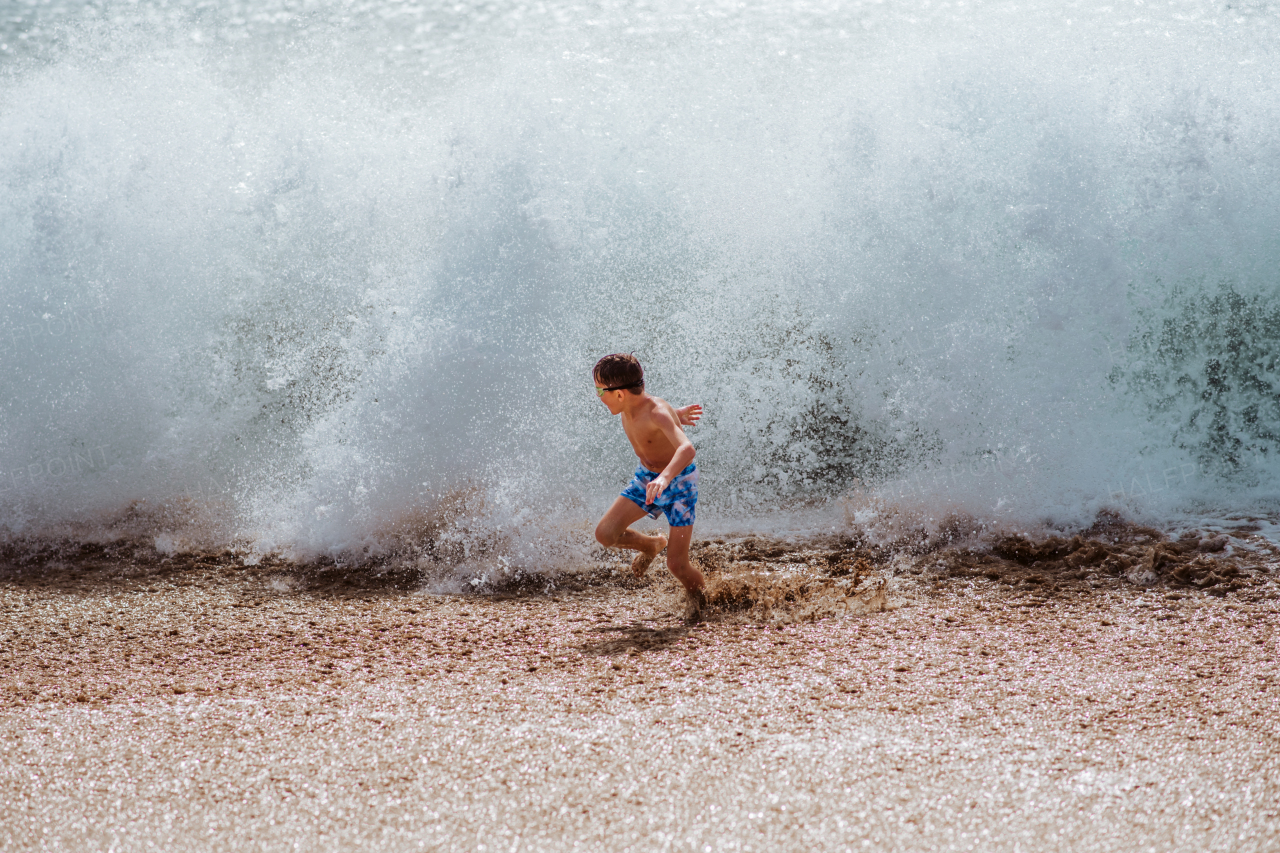 Young boy playing, running and splashing in strong sea waves. Smilling boy in swimsuit and swimming goggles. Concept of a beach summer vacation with kids.