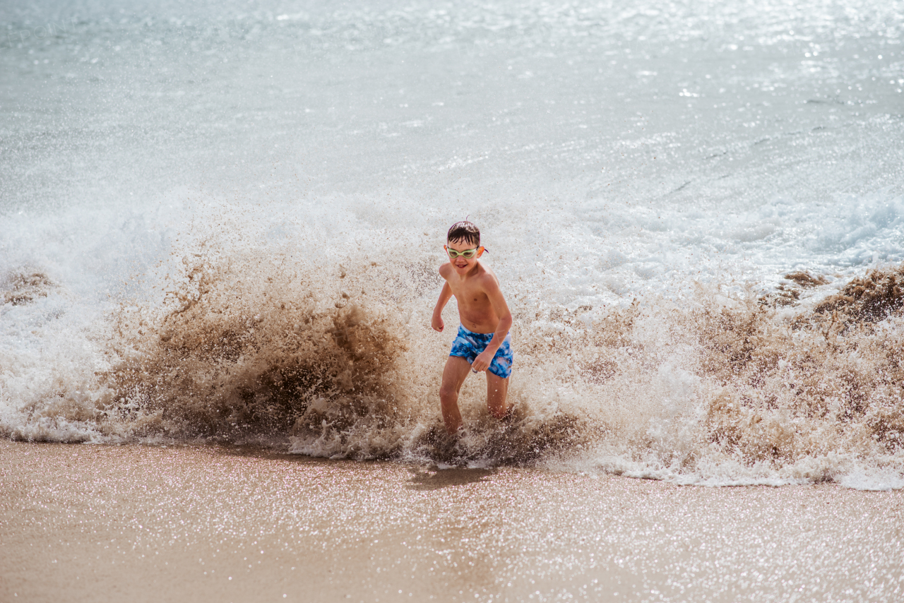 Young boy playing, running and splashing in strong sea waves. Smilling boy in swimsuit looking at waves. Concept of a beach summer vacation with kids.