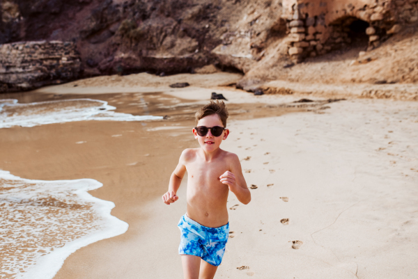 Young boy playing, running and splashing in strong sea waves. Smilling boy in swimsuit and swimming goggles. Concept of a beach summer vacation with kids.