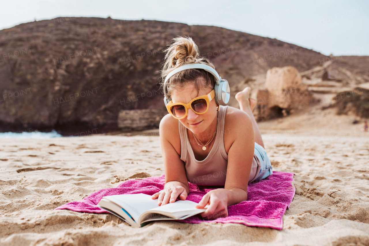 Woman on vacation, lying on towel at beach, reading book and listening music via headphones. Concept of a beach summer vacation.