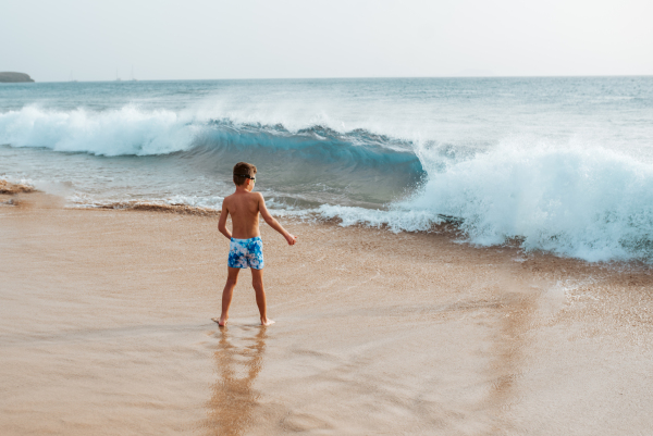 Young boy playing, running and splashing in strong sea waves. Smilling boy in swimsuit looking at waves. Concept of a beach summer vacation with kids.