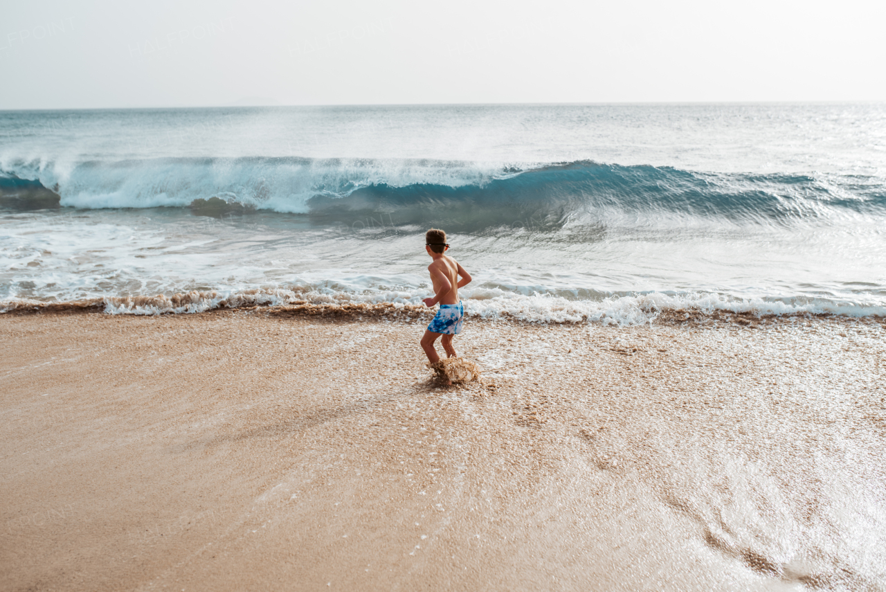 Young boy playing, running and splashing in strong sea waves. Smilling boy in swimsuit looking at waves. Concept of a beach summer vacation with kids.