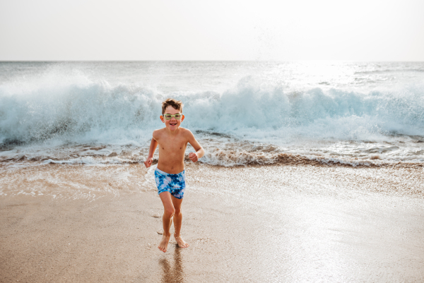 Young boy playing, running and splashing in strong sea waves. Smilling boy in swimsuit and swimming goggles. Concept of a beach summer vacation with kids.