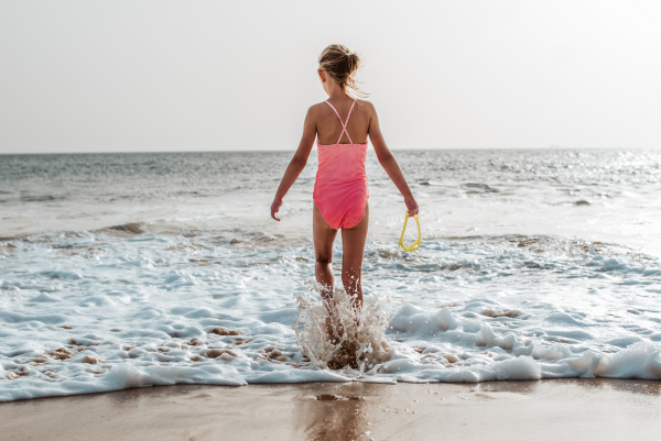 Young blonde girl standing on beach in swimsuit, holding yellow swimming goggles in hand, rear view. Canary Islands. Concept of beach summer vacation with kids.