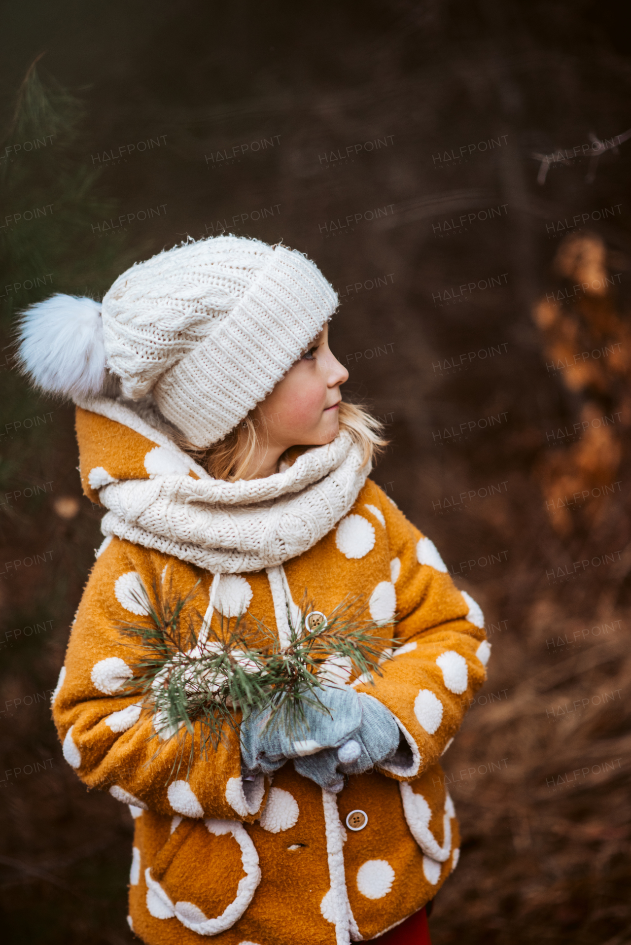 Side view of a little girl standing in the forest, dressed in warm clothing, holding pine needles. Spending time outdoors, early spring, autumn season.