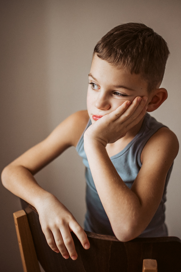 The boy is bored, sitting on a chair and resting his head on his hand.