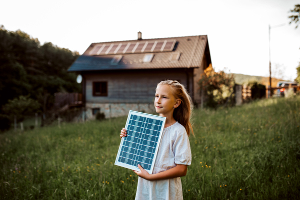 Little girl with model of solar panel, standing in the middle of meadow, house with solar panels behind. Concept of renewable resources, sustainability and green energy.