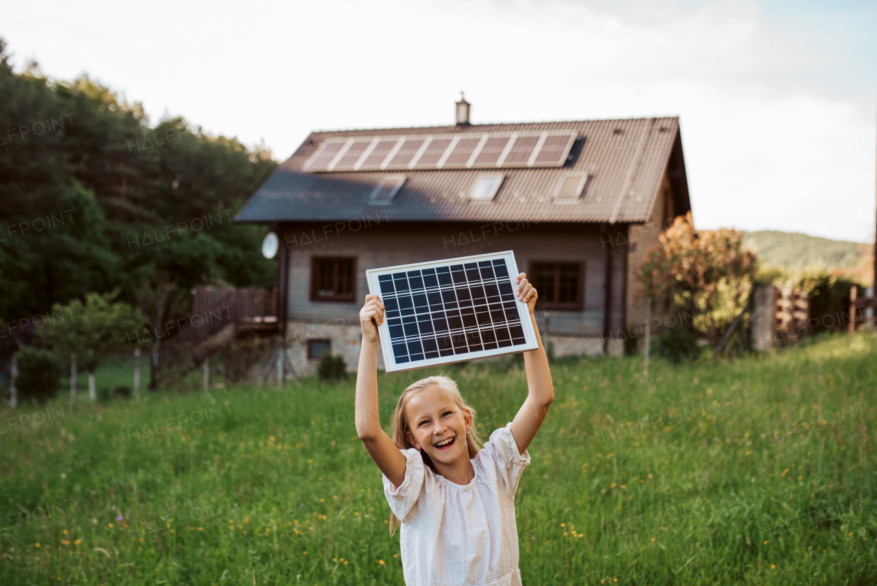 Little girl with model of solar panel, standing in the middle of meadow, house with solar panels behind. Concept of renewable resources, sustainability and green energy.