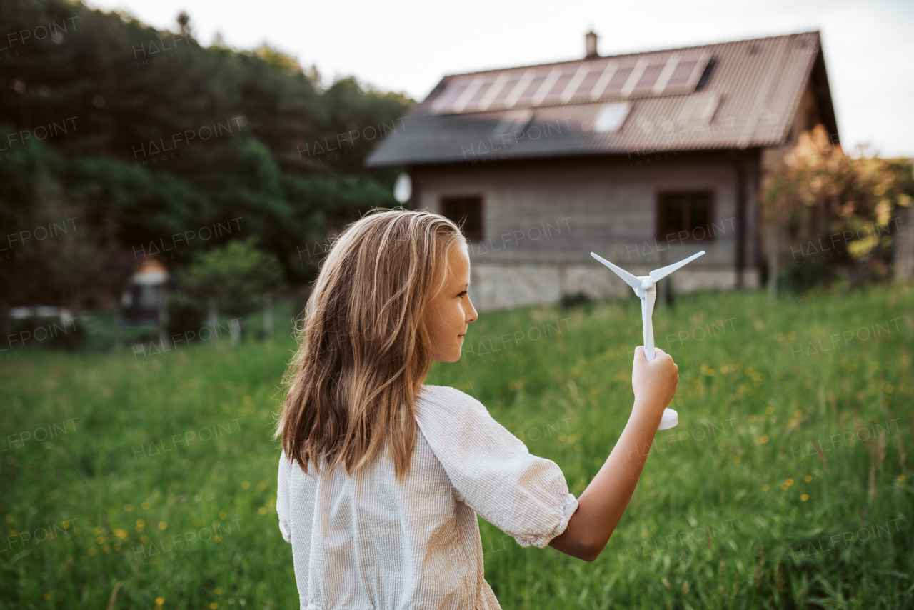 Girl with model of wind turbine, standing in the middle of meadow, house with solar panels behind. Concept of renewable resources, sustainability and green energy.