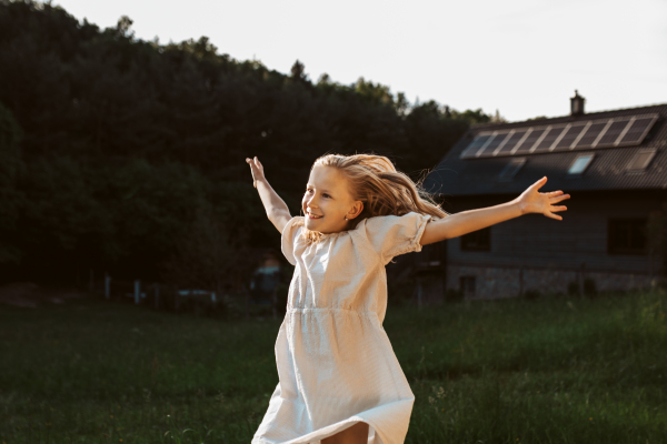 Little girl in front of house with solar panels on roof, jumping, dancing and having fun. Concept of renewable resources, sustainability and green energy.