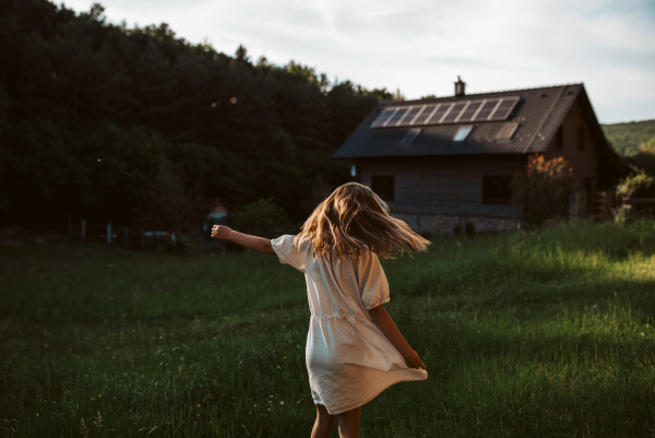 Little girl in front of house with solar panels on roof, jumping, dancing and having fun. Concept of renewable resources, sustainability and green energy.