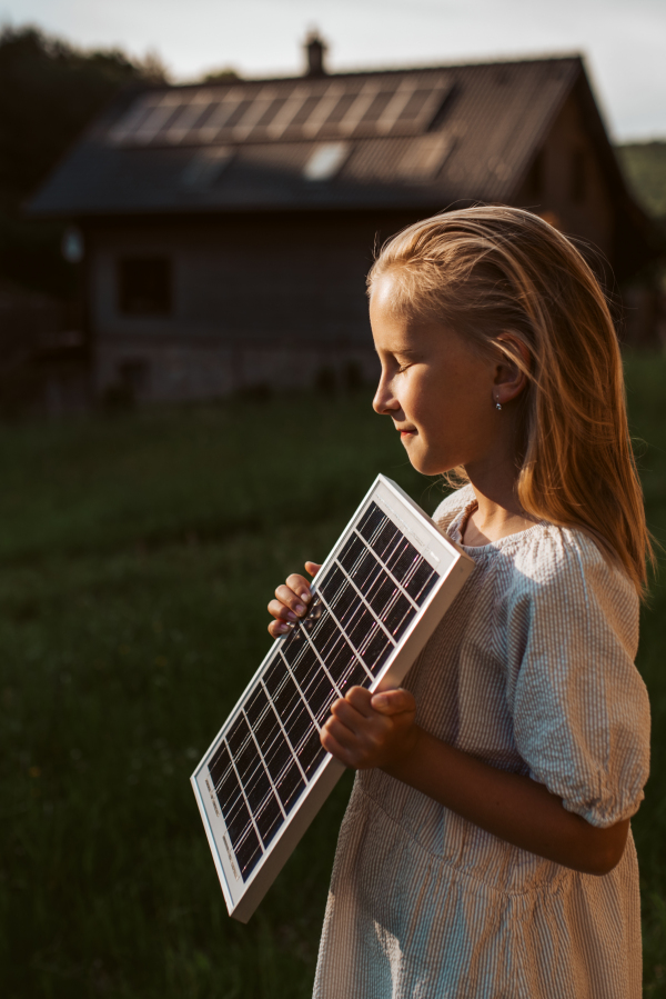 Little girl with model of solar panel, standing in the middle of meadow, house with solar panels behind. Concept of renewable resources, sustainability and green energy.