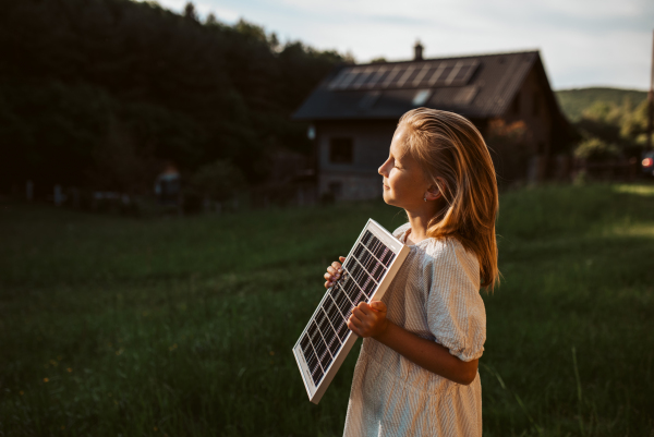 Little girl with model of solar panel, standing in the middle of meadow, house with solar panels behind. Concept of renewable resources, sustainability and green energy.