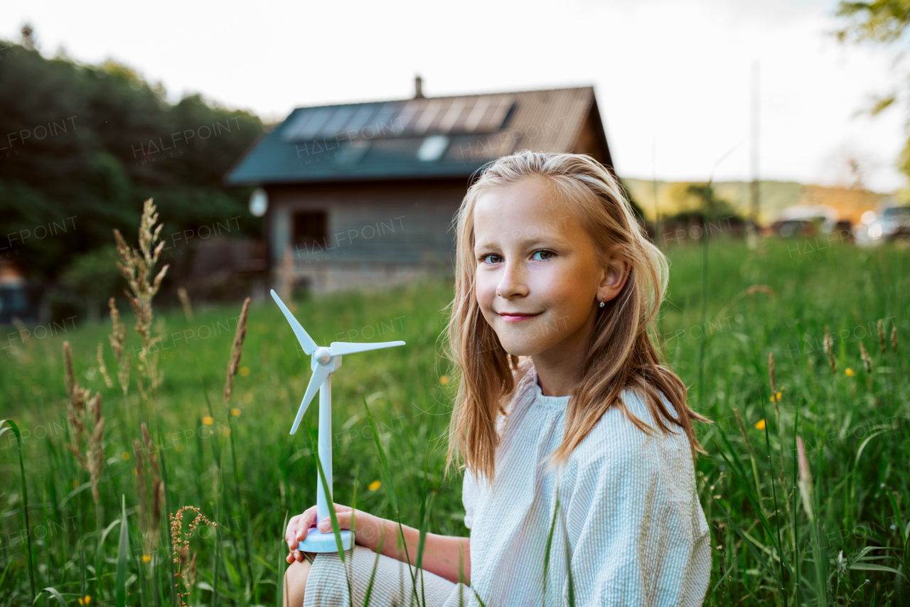 Girl with model of wind turbine, standing in the middle of meadow, house with solar panels behind. Concept of renewable resources, sustainability and green energy.