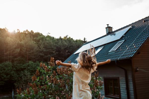 Little girl in front of house with solar panels on roof, jumping, dancing and having fun. Concept of renewable resources, sustainability and green energy.