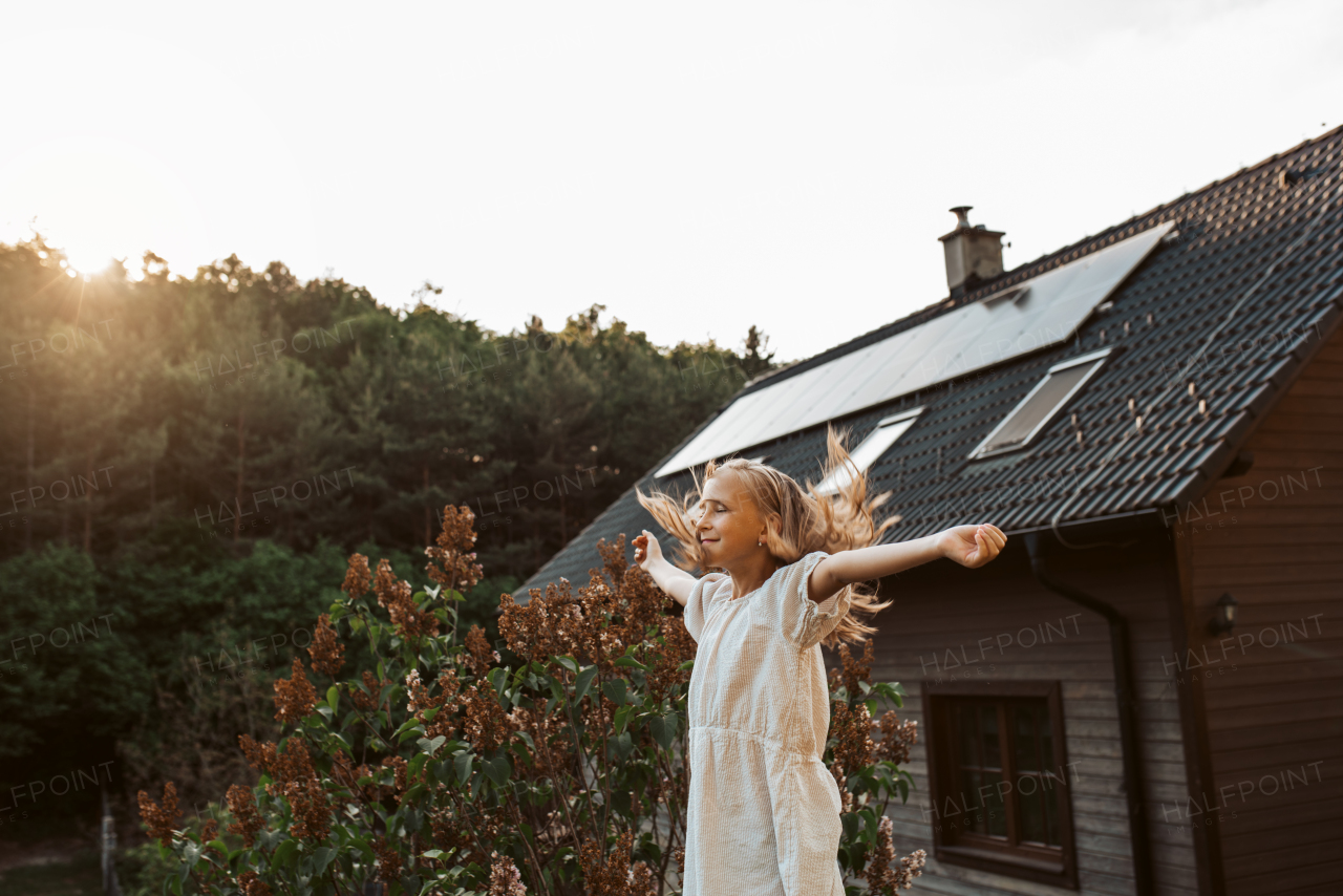 Little girl in front of house with solar panels on roof, eyes closed, standing with open arms. Concept of renewable resources, sustainability and green energy.