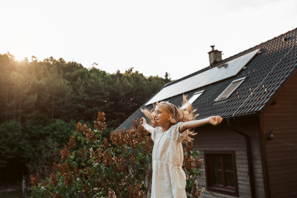 Little girl in front of house with solar panels on roof, eyes closed, standing with open arms. Concept of renewable resources, sustainability and green energy.