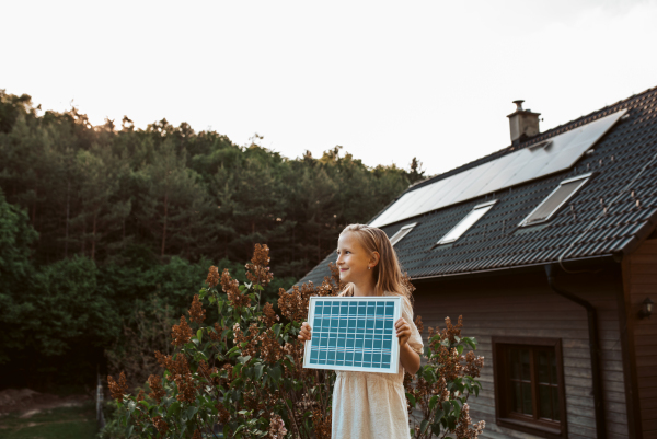 Little girl with model of solar panel, standing in the middle of meadow, house with solar panels behind. Concept of renewable resources, sustainability and green energy.