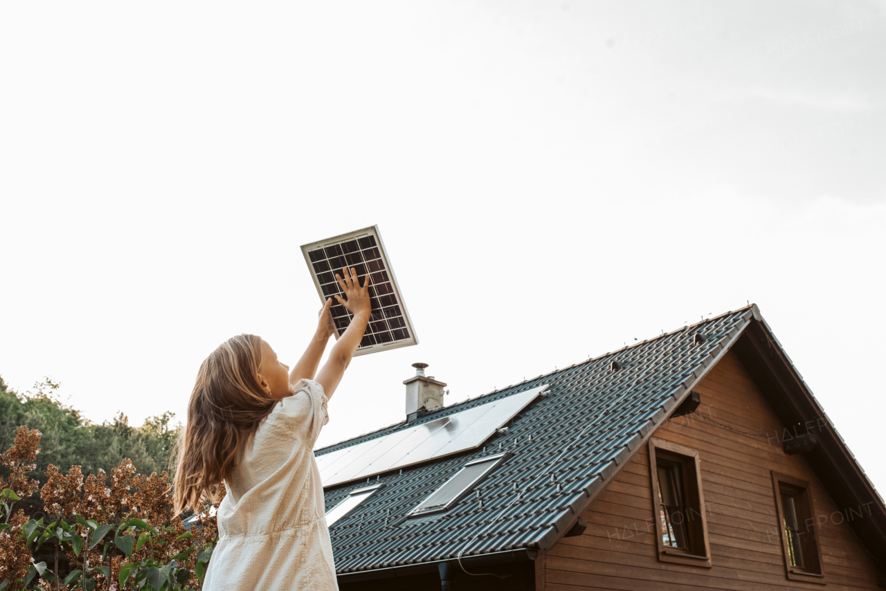 Little girl with model of solar panel, standing in the middle of meadow, house with solar panels behind. Concept of renewable resources, sustainability and green energy.